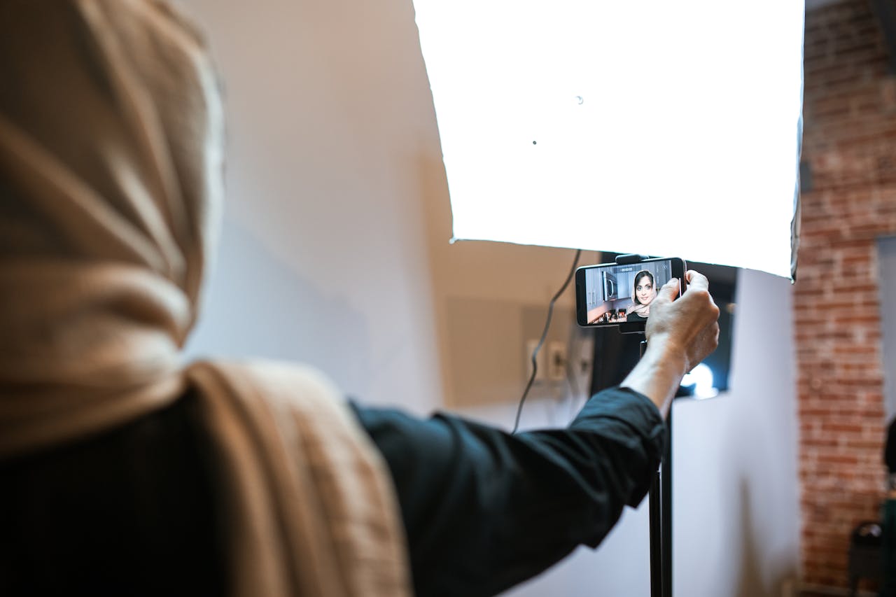 A woman wearing a headscarf is recording a vlog with a smartphone and bright studio light.
